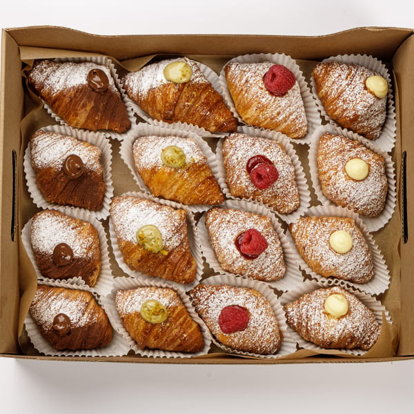 A box of mini sfogliatelle and lobster-tail pastries dusted with powdered sugar and topped with cream, raspberries, and pistachio.