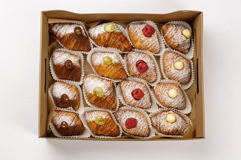 A box of mini sfogliatelle and lobster-tail pastries dusted with powdered sugar and topped with cream, raspberries, and pistachio.