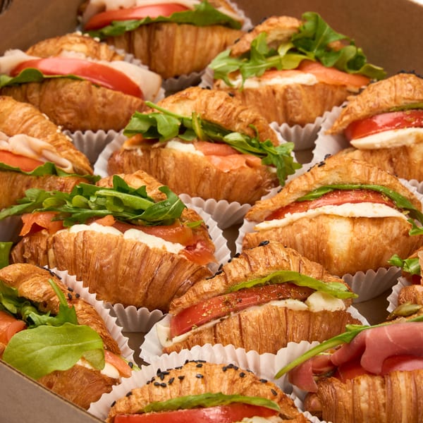 A box filled with various mini croissant sandwiches, including turkey, salmon, and prosciutto.