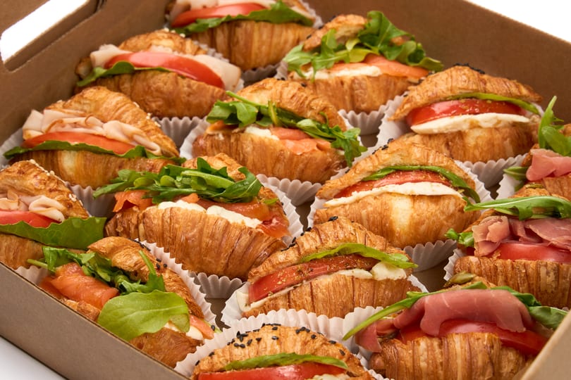 A box filled with various mini croissant sandwiches, including turkey, salmon, and prosciutto.