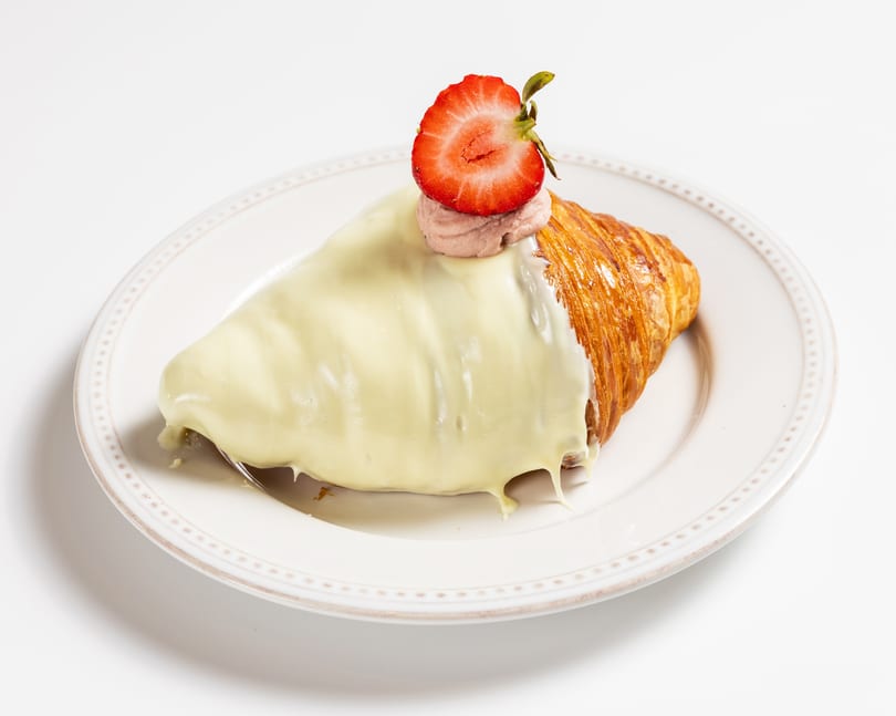 Croissant covered in white chocolate, topped with strawberry cream and a fresh strawberry half.