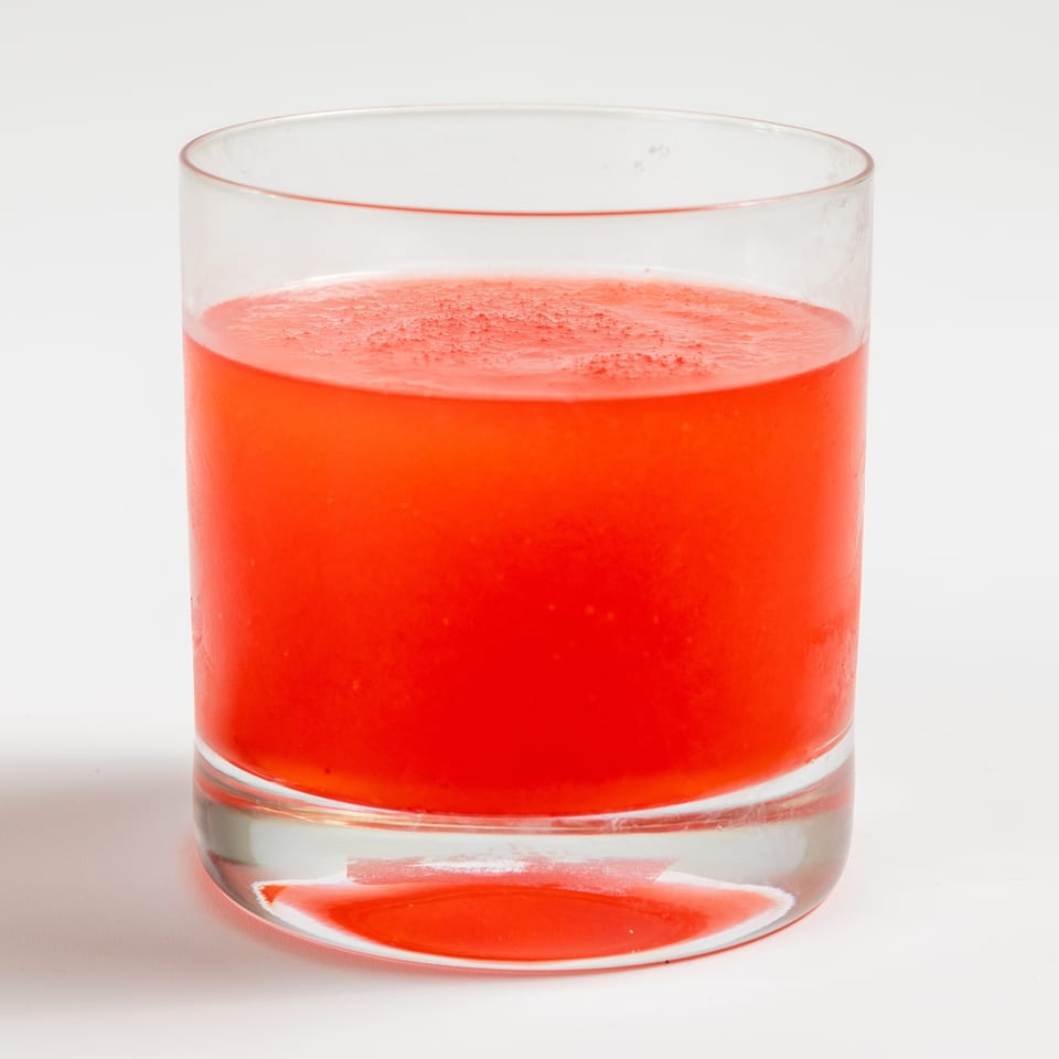 A clear glass filled with a vibrant red beverage, possibly a cocktail or juice, on a white background.