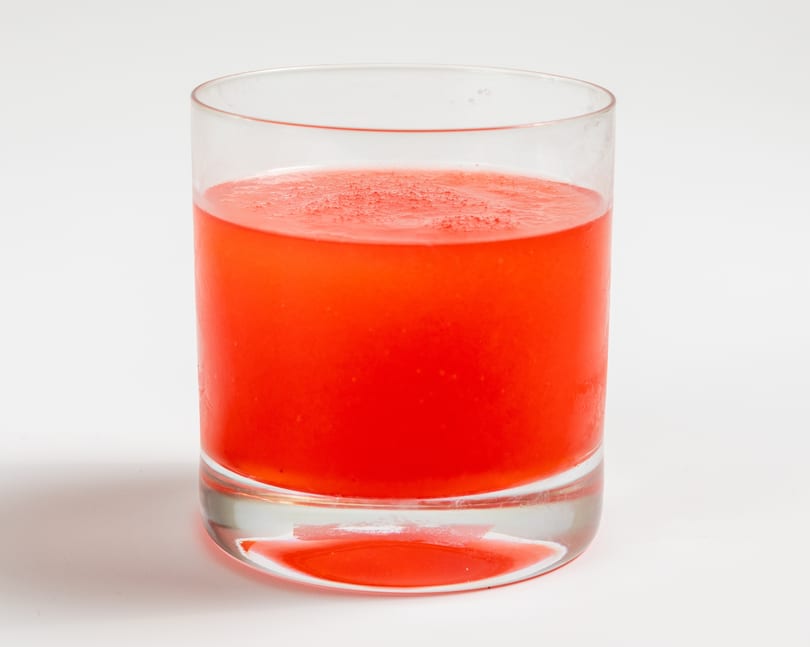 A clear glass filled with a vibrant red beverage, possibly a cocktail or juice, on a white background.