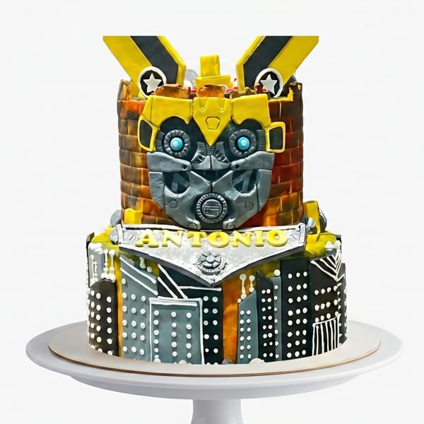 Two-tiered Transformers Bumblebee cake with "Antonio" written on the front, on a white cake stand.