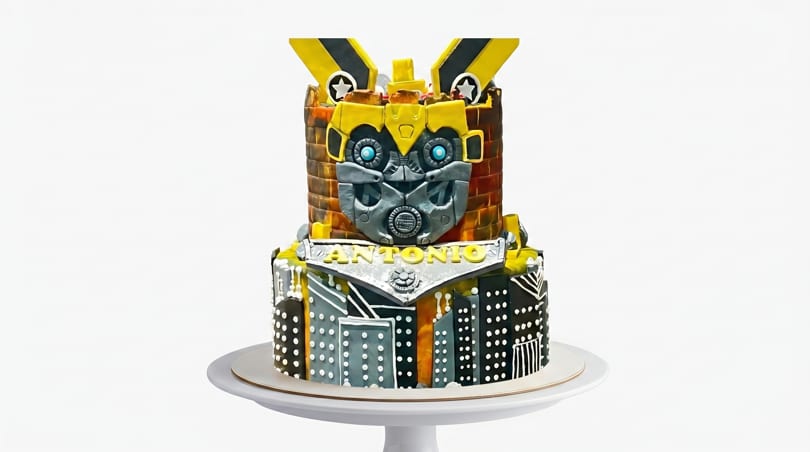 Two-tiered Transformers Bumblebee cake with "Antonio" written on the front, on a white cake stand.