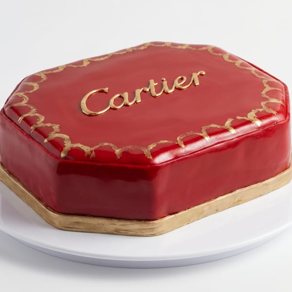 A red, octagonal cake with gold trim and "Cartier" written in gold on top, on a white plate.