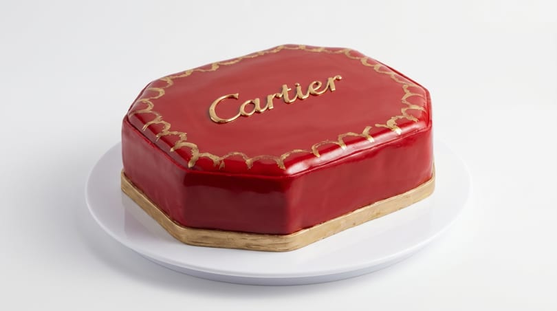 A red, octagonal cake with gold trim and "Cartier" written in gold on top, on a white plate.