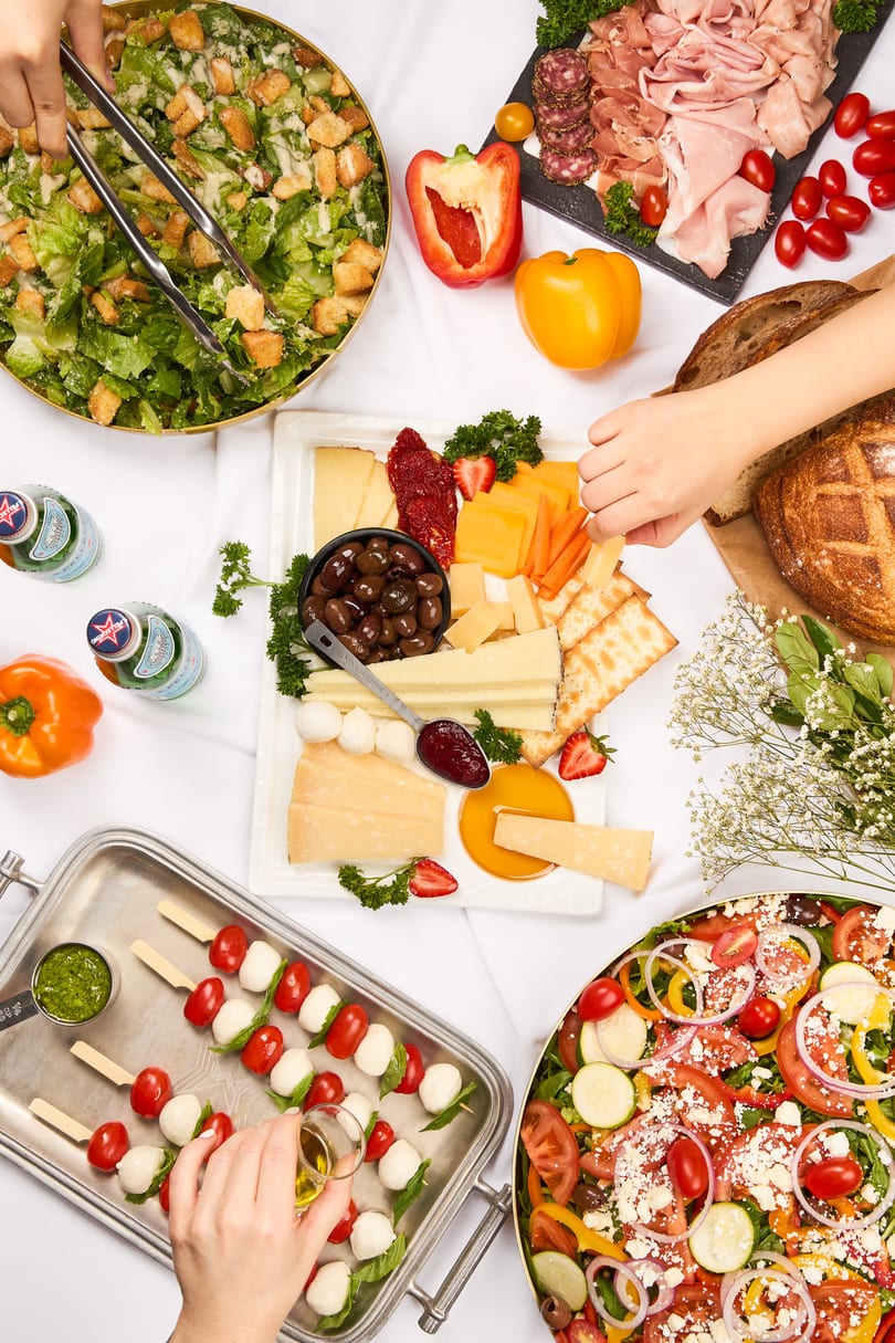 Overhead view of a buffet spread with salads, charcuterie, cheese board, and caprese skewers.
