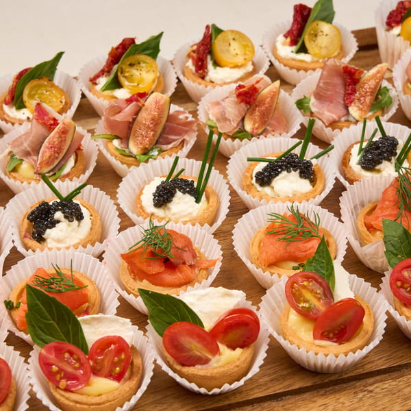 A wooden tray filled with rows of assorted mini tarts, including caviar, salmon, prosciutto, and caprese.