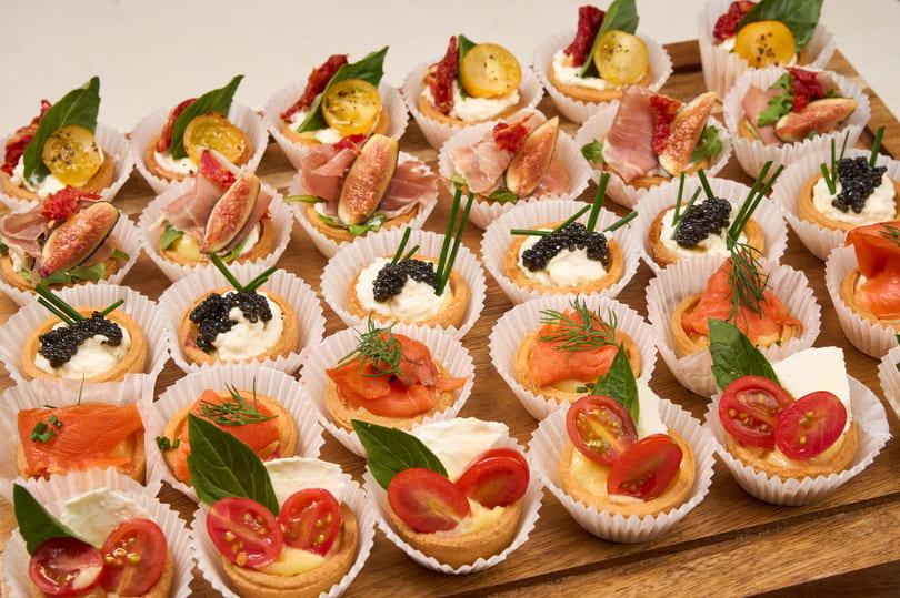 A wooden tray filled with rows of assorted mini tarts, including caviar, salmon, prosciutto, and caprese.