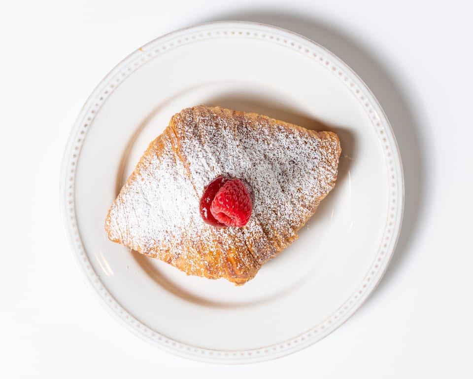 Golden croissant dusted with powdered sugar, topped with a fresh raspberry and jam on a white plate.