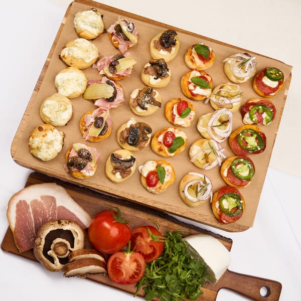 Assortment of mini pizzas and bruschetta on a wooden board, with fresh ingredients below.