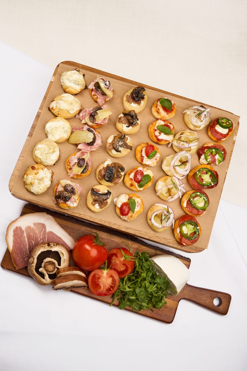 Assortment of mini pizzas and bruschetta on a wooden board, with fresh ingredients below.