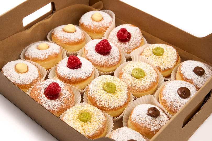 A box of powdered sugar donuts with various toppings including raspberries, yellow cream, and green cream.