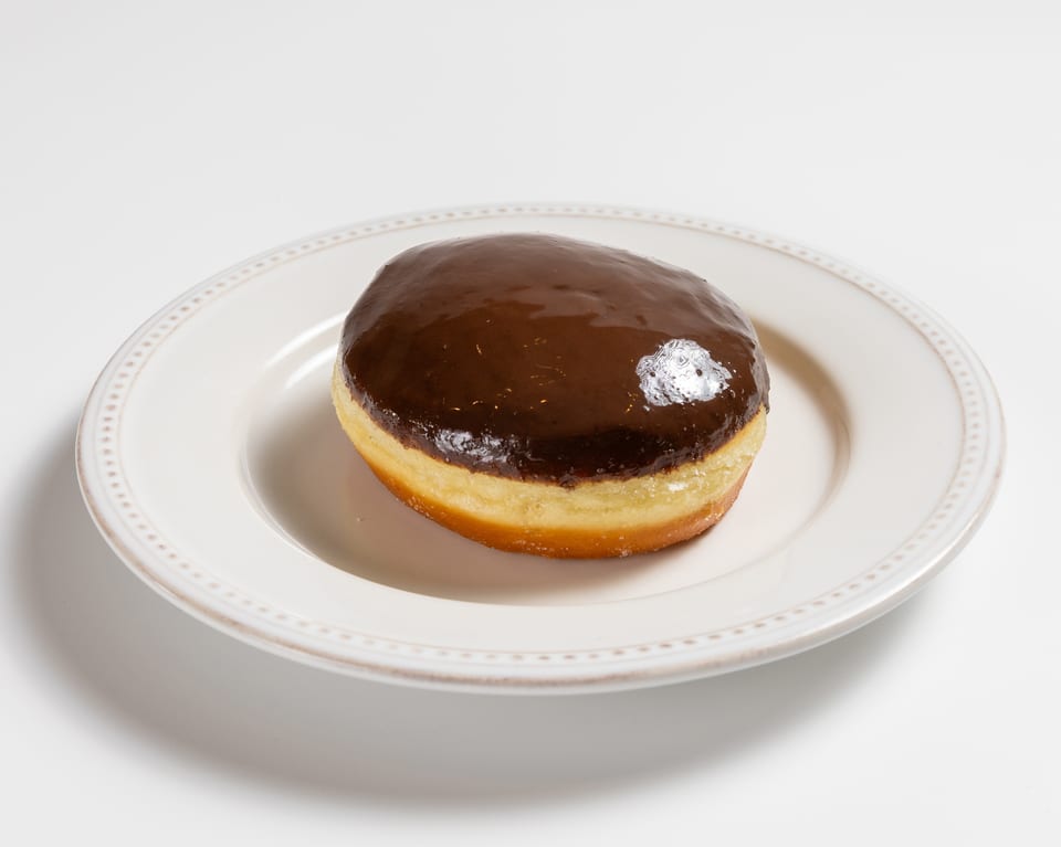 A chocolate-glazed donut on a white plate with a decorative rim, against a white background.