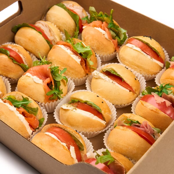 A catering box filled with various mini sandwiches, including salmon, prosciutto, and caprese.