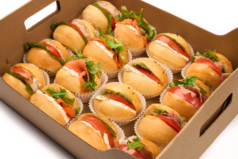 A catering box filled with various mini sandwiches, including salmon, prosciutto, and caprese.
