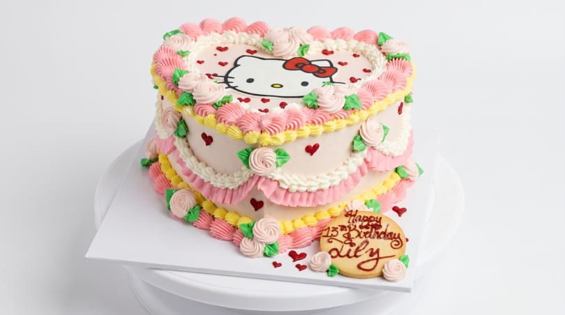 Heart-shaped Hello Kitty cake with pink, yellow, and white frosting, roses, and red hearts.