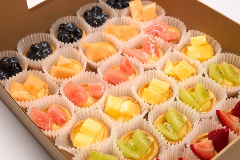 A cardboard box filled with rows of mini fruit tarts, each in a white paper cup.
