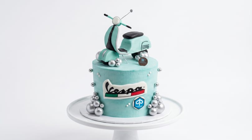 A light blue Vespa-themed cake with a miniature scooter on top and silver edible pearls.