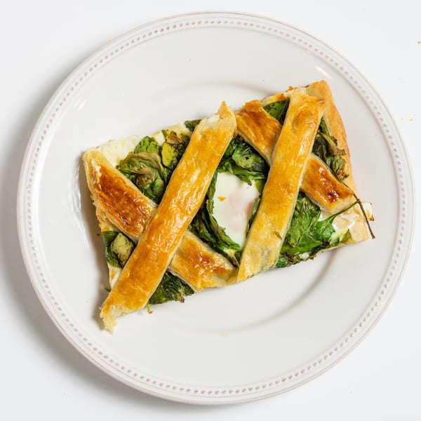 A slice of savory tart with a lattice crust, spinach, and a baked egg on a white plate.