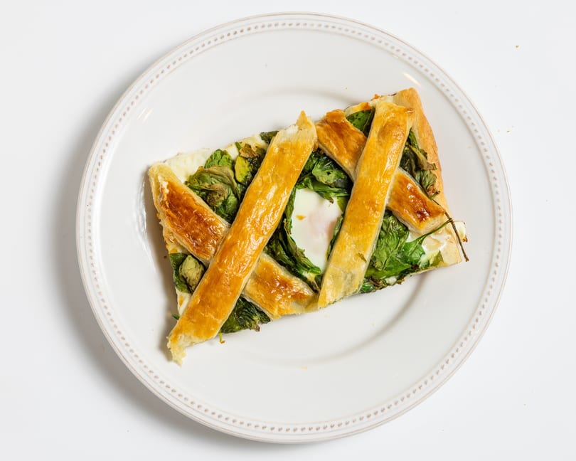 A slice of savory tart with a lattice crust, spinach, and a baked egg on a white plate.