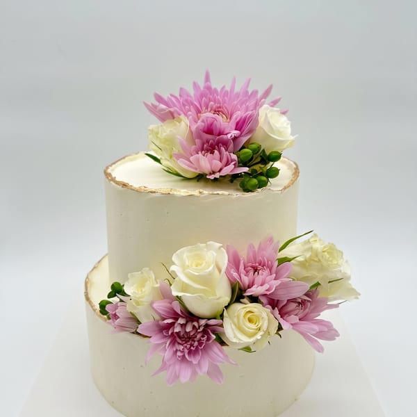 A two-tiered white buttercream cake with gold trim, topped with white roses and pink chrysanthemums.