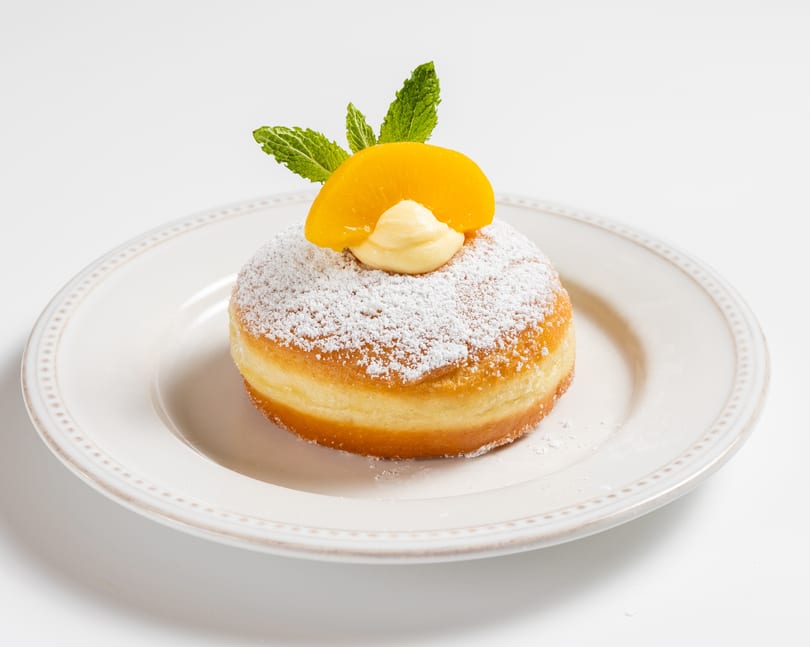 Powdered sugar donut with cream, a peach slice, and mint on a white plate.