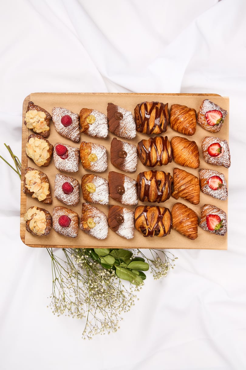 Assortment of croissants with various toppings like almonds, raspberries, chocolate, and strawberries on a wooden board.