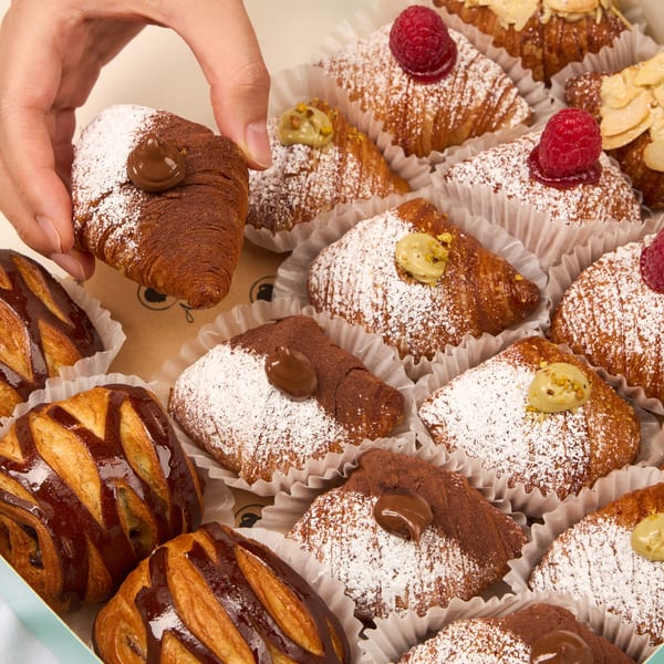 Hand picking a chocolate croissant from a box of assorted pastries with raspberries, almonds, and pistachios.