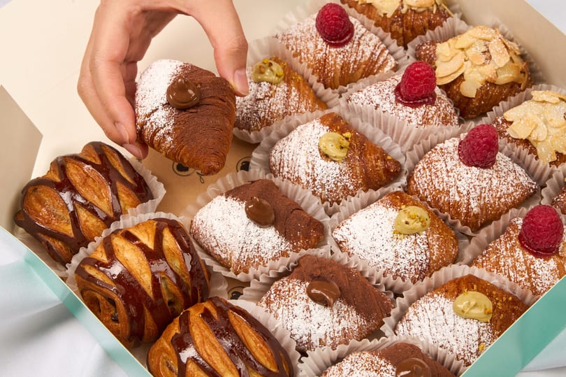 Hand picking a chocolate croissant from a box of assorted pastries with raspberries, almonds, and pistachios.