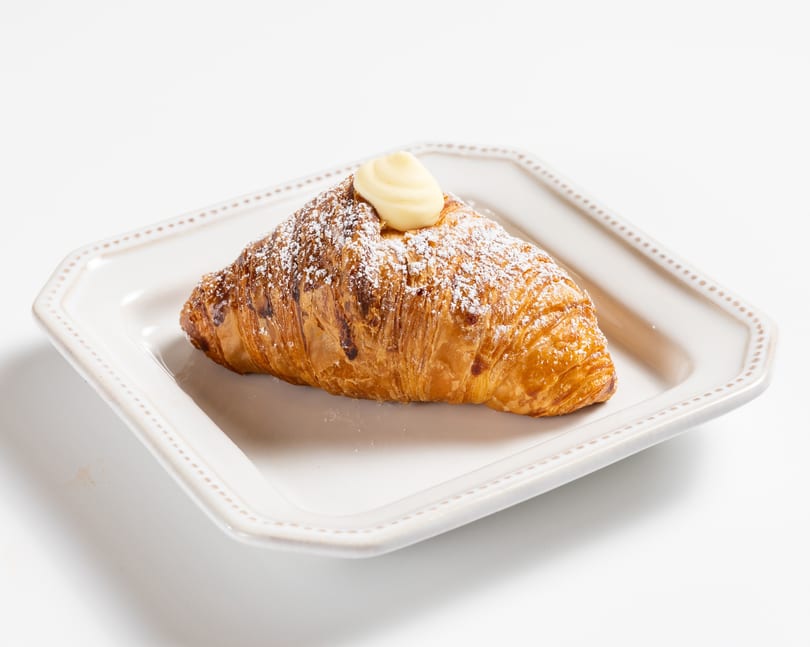 Golden croissant dusted with powdered sugar and topped with a dollop of cream on a white plate.