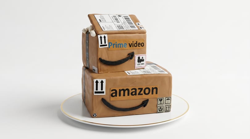 A two-tiered cake designed as stacked Amazon Prime boxes, on a white plate with a gold rim.