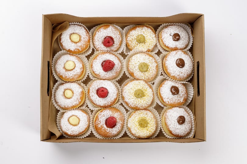 A box of mini bomboloni donuts dusted with powdered sugar and filled with cream, raspberry, pistachio, and chocolate.