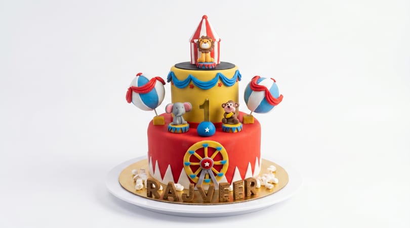 A vibrant, two-tiered circus-themed birthday cake with animal figurines, a Ferris wheel, and the name "RAJVEER".