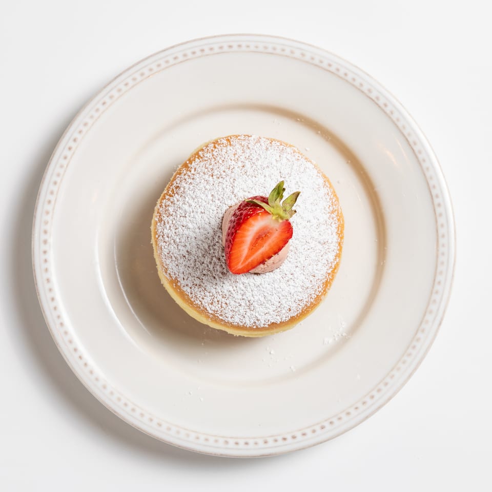 A fluffy pancake dusted with powdered sugar, topped with a dollop of cream and a fresh strawberry half.