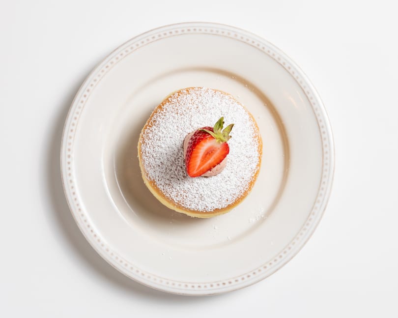 A fluffy pancake dusted with powdered sugar, topped with a dollop of cream and a fresh strawberry half.