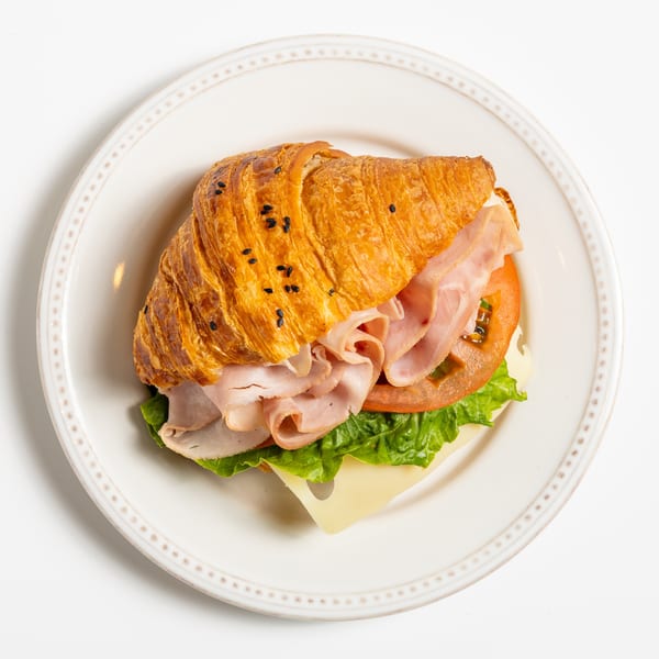 A croissant sandwich with ham, cheese, lettuce, and tomato on a white plate.