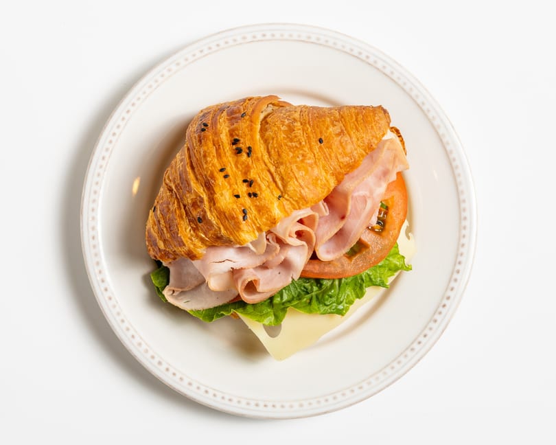 A croissant sandwich with ham, cheese, lettuce, and tomato on a white plate.