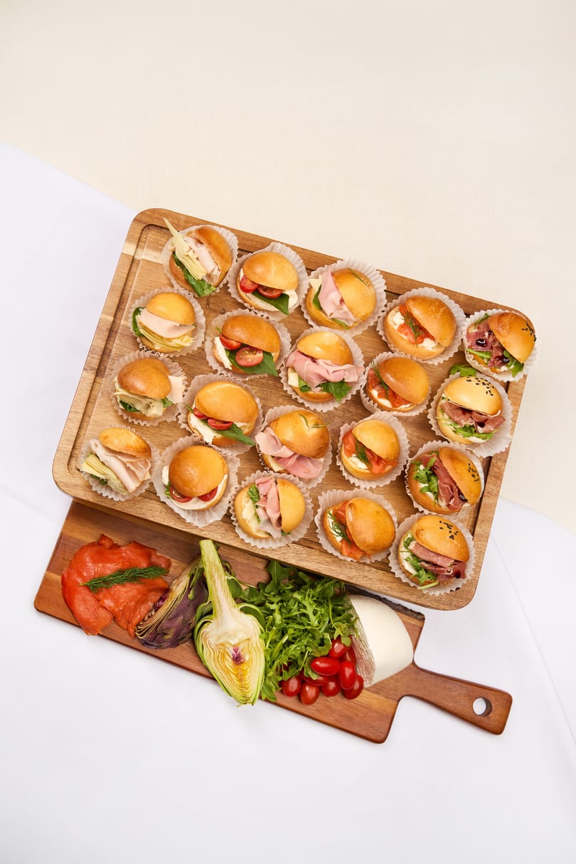 Assortment of mini sandwiches on wooden boards with fresh ingredients like salmon, artichoke, and mozzarella.
