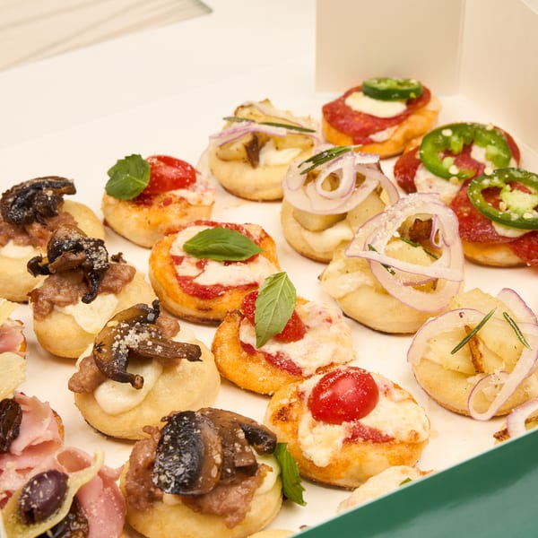A box of assorted baby pizzas topped with pepperoni, jalapeno, cherry tomato, mushroom, and caramelized onion.
