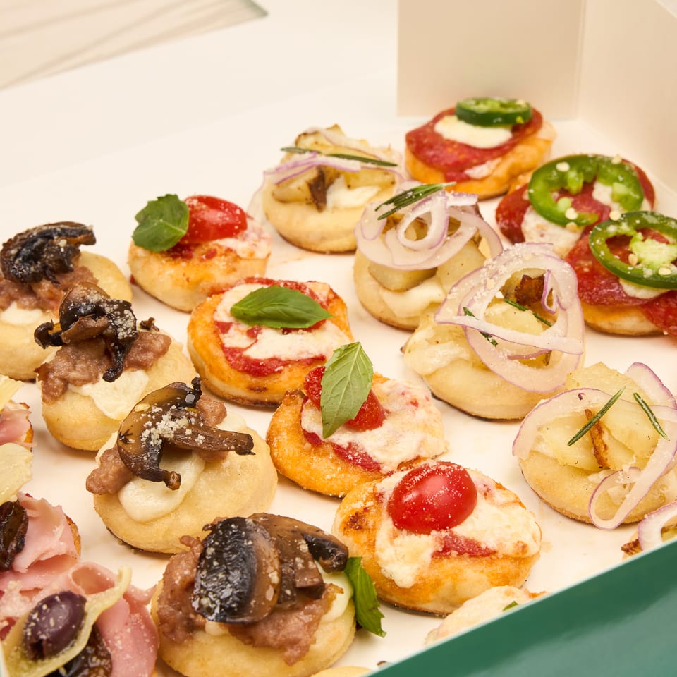 A box of assorted baby pizzas topped with pepperoni, jalapeno, cherry tomato, mushroom, and caramelized onion.