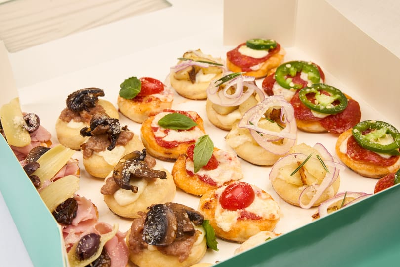 A box of assorted baby pizzas topped with pepperoni, jalapeno, cherry tomato, mushroom, and caramelized onion.