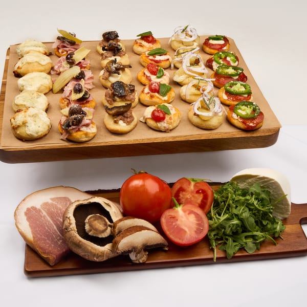 A wooden board of assorted baby pizzas with fresh mozzarella, tomato, prosciutto, and fresh ingredients alongside.