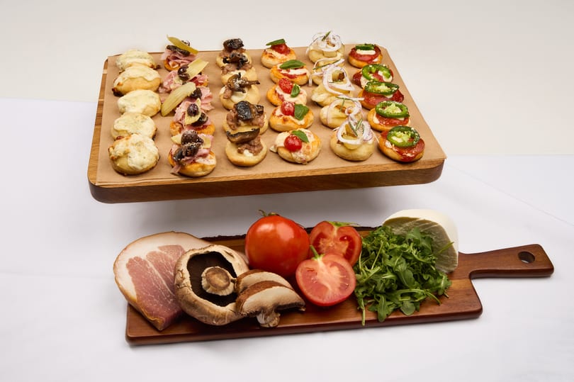 A wooden board of assorted baby pizzas with fresh mozzarella, tomato, prosciutto, and fresh ingredients alongside.