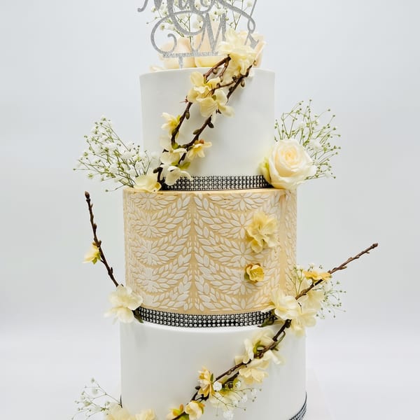 A three-tiered white and gold wedding cake with a 'Mr & Mrs' topper, cream roses, blossoms, and crystal banding.