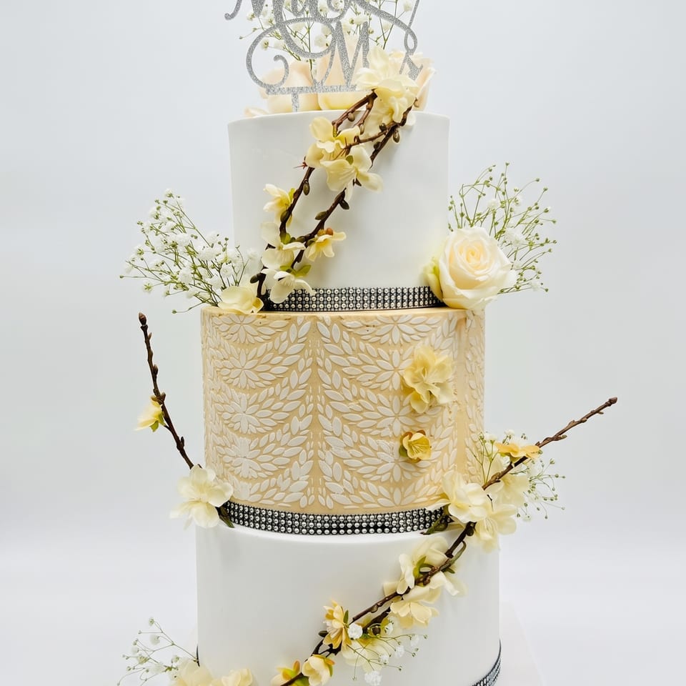 A three-tiered white and gold wedding cake with a 'Mr & Mrs' topper, cream roses, blossoms, and crystal banding.