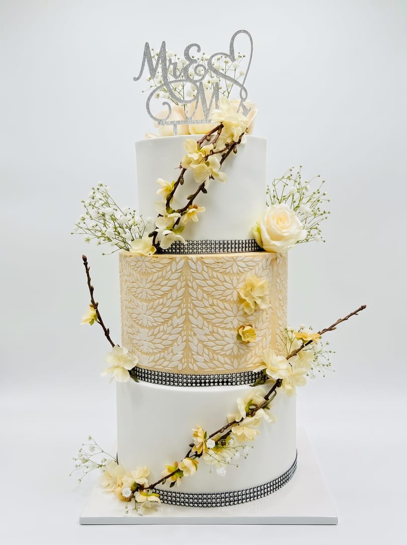 A three-tiered white and gold wedding cake with a 'Mr & Mrs' topper, cream roses, blossoms, and crystal banding.