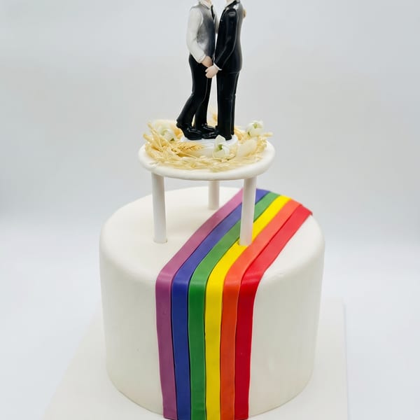 A white wedding cake with a rainbow fondant stripe and two-groom cake topper surrounded by wheat and calla lilies.