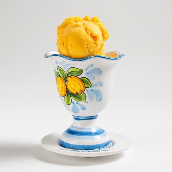A scoop of bright yellow gelato in a decorative white cup with blue and yellow lemon designs.